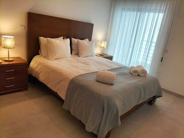 A neatly made bed with white linens, a grey throw blanket, and two rolled towels on the foot of the bed. There are two bedside tables with lamps, one on each side of the bed.