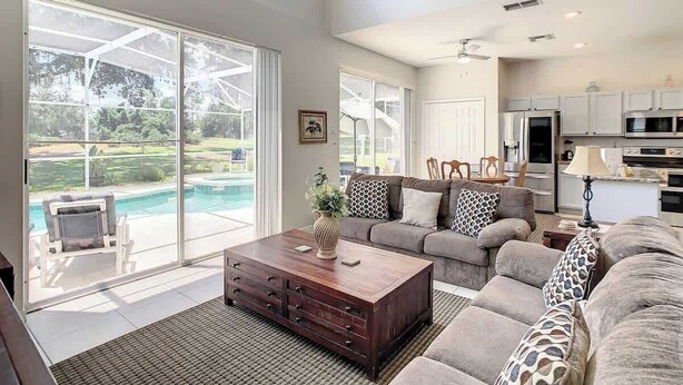 A spacious living room with a large sliding glass door leading to a pool, a cozy seating area with a sofa and a coffee table, and a kitchen in the background. - Spacious Getaway with Pool on stunning Golf Course (Haines City)