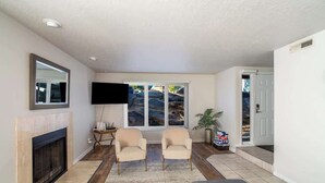 A living room with a fireplace, two armchairs, a TV, and a large window with a view of hills. - Pet-Friendly Condo with Private Hot Tub & Views (St. George)