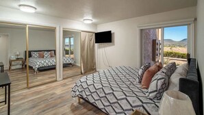 A bedroom with a large bed, a TV mounted on the wall, and a sliding glass door leading to a balcony with a view of the hills. - Pet-Friendly Condo with Private Hot Tub & Views (St. George)