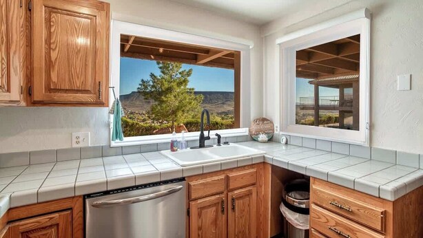 A kitchen with wooden cabinets, a stainless steel dishwasher, a window with a view of a tree and landscape, and a tiled countertop. - Pet-Friendly Condo with Private Hot Tub & Views (St. George)