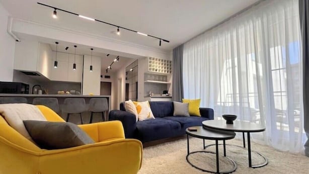 Ein modernes Wohnzimmer mit Sofa, Sessel und Couchtisch. Im Hintergrund ist eine Küche mit Schränken und einem Essbereich zu sehen.