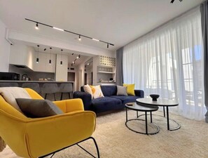 A modern living room with a sofa, armchair, and coffee table. There is a kitchen in the background with cabinets and a dining area. - The First House Free Parking (Timișoara)