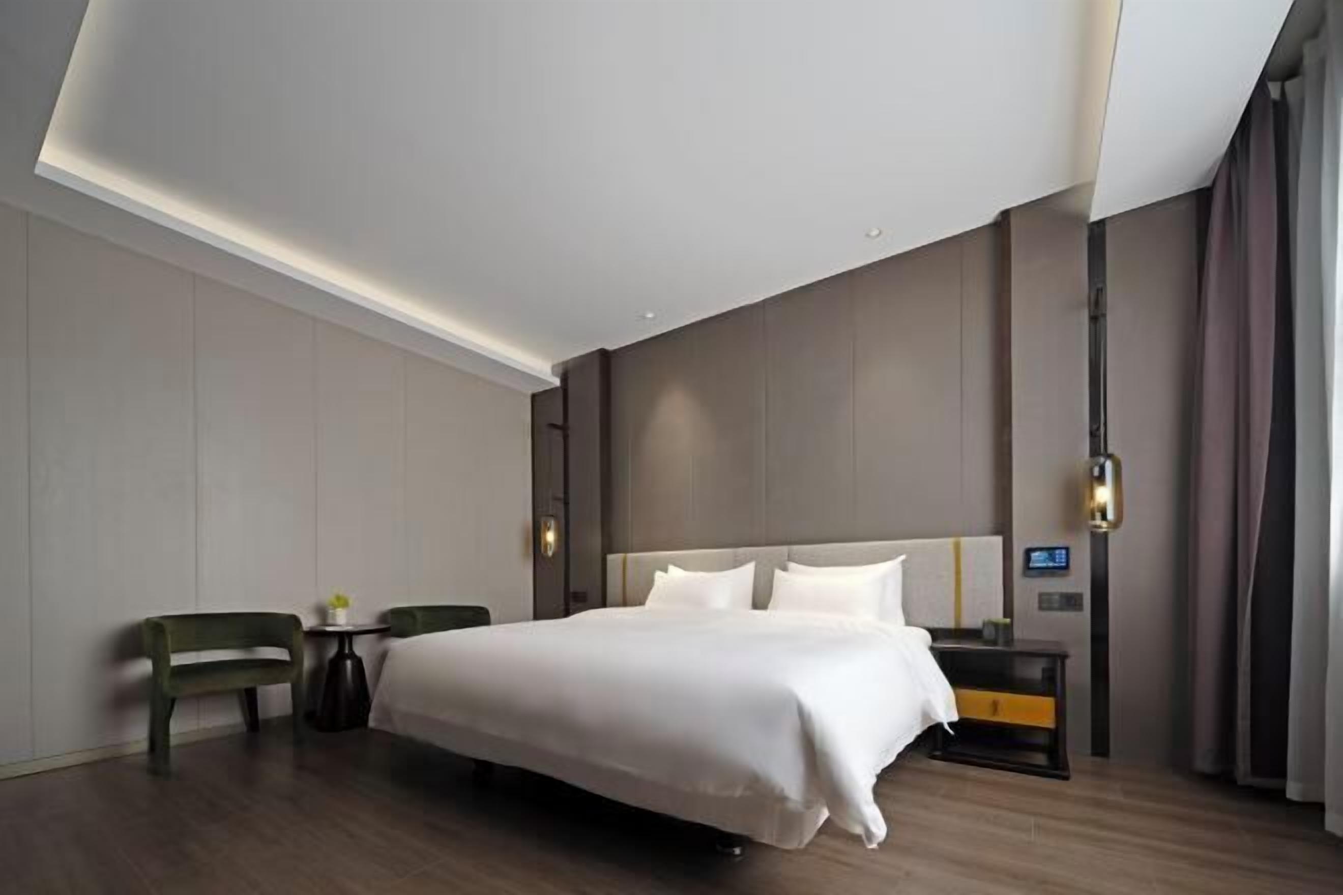 A modern hotel room with a large bed, a bedside table, a chair, and a small table with a vase.
