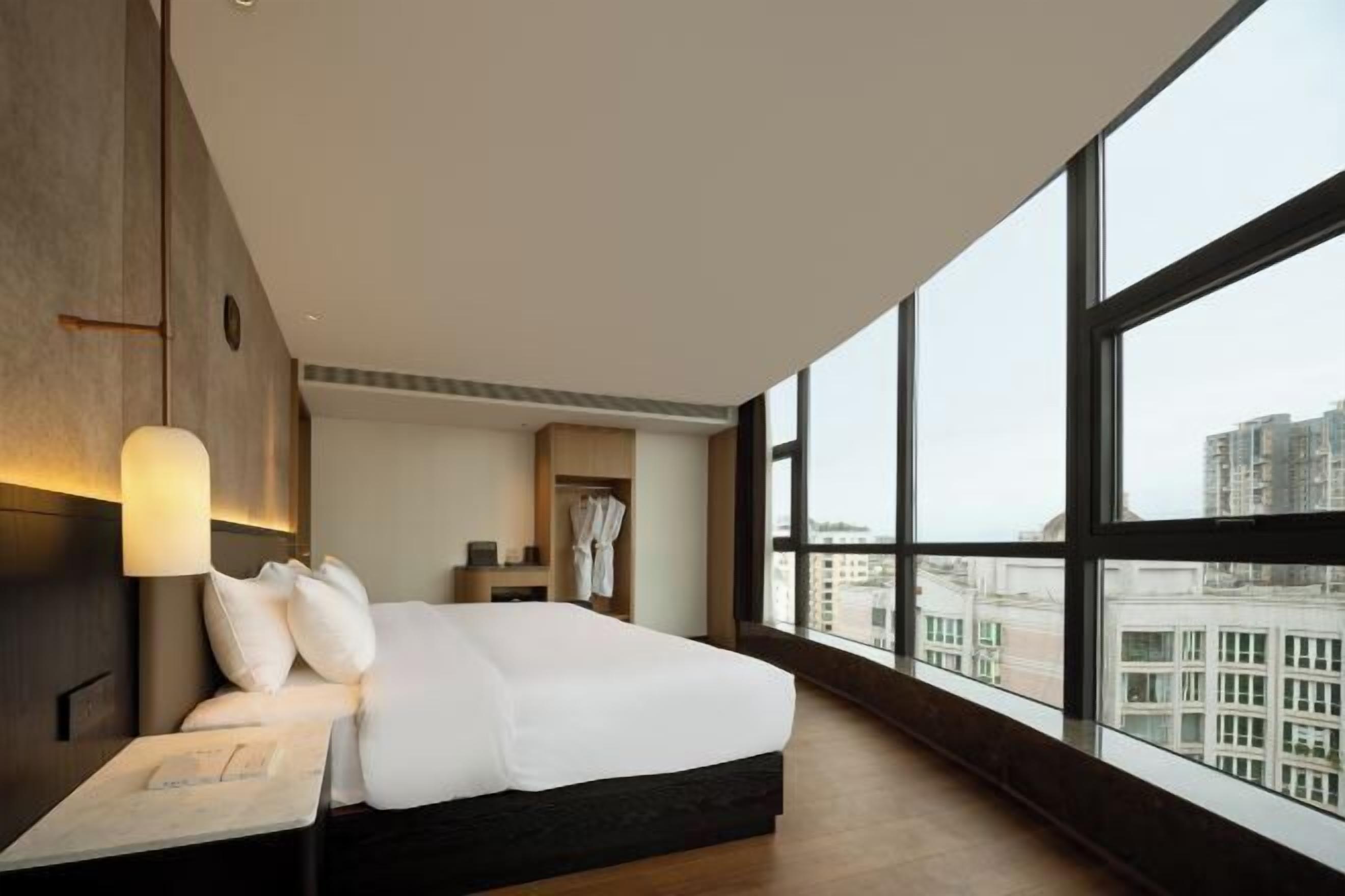 Quarto de hotel moderno com uma cama grande, uma mesa de cabeceira, uma luminária, uma janela com vista para a cidade e um guarda-roupa.