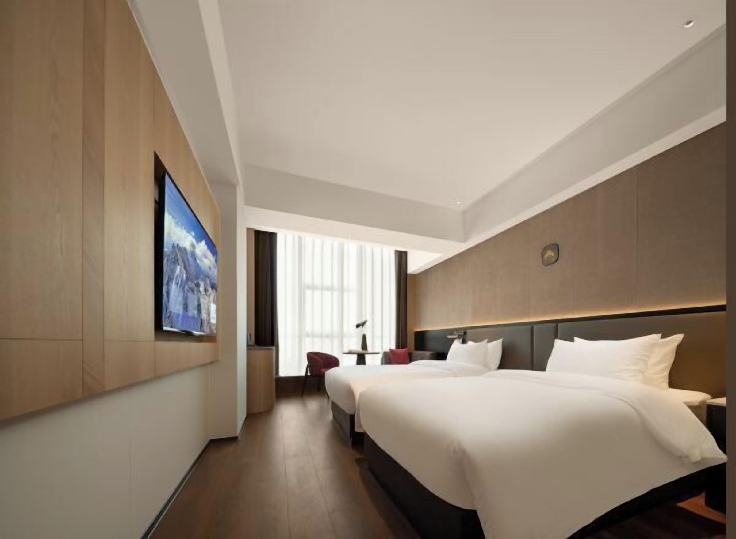 A hotel room with two beds, a TV mounted on the wall, a large window, and a clock on the wall.