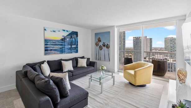 A modern living room with a dark grey sofa, a glass coffee table, a yellow armchair, and a large window with a city view.