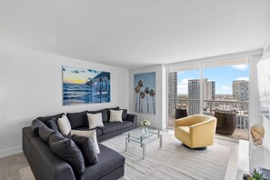 A modern living room with a dark grey sofa, a glass coffee table, a yellow armchair, and a large window with a city view. - Spacious 3 bedrooms with Parking (Miami)