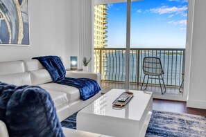 Een moderne woonkamer met een witte bank, een blauwe plaid en een bijzettafeltje. Een groot raam biedt uitzicht op een stadshemelbeeld en een wateroppervlak.