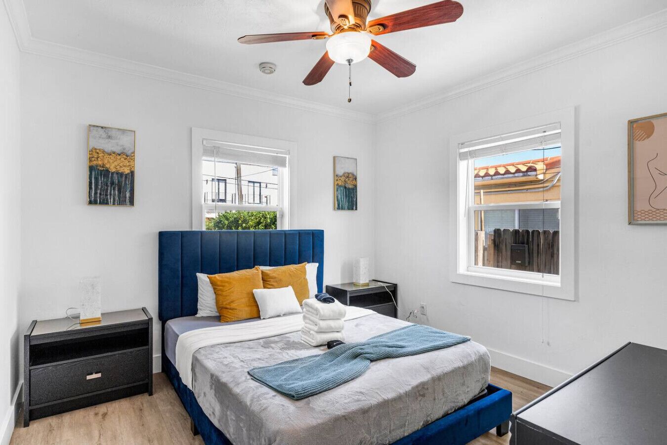 A bedroom with a bed, a nightstand, a desk, a ceiling fan, and artwork on the walls.