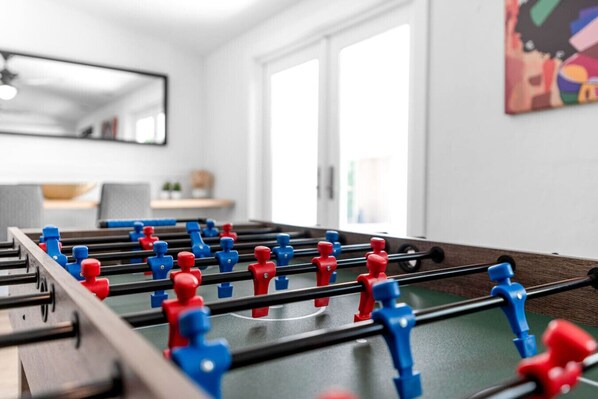 A foosball table in a room with a large window, a mirror on the wall, and a painting.