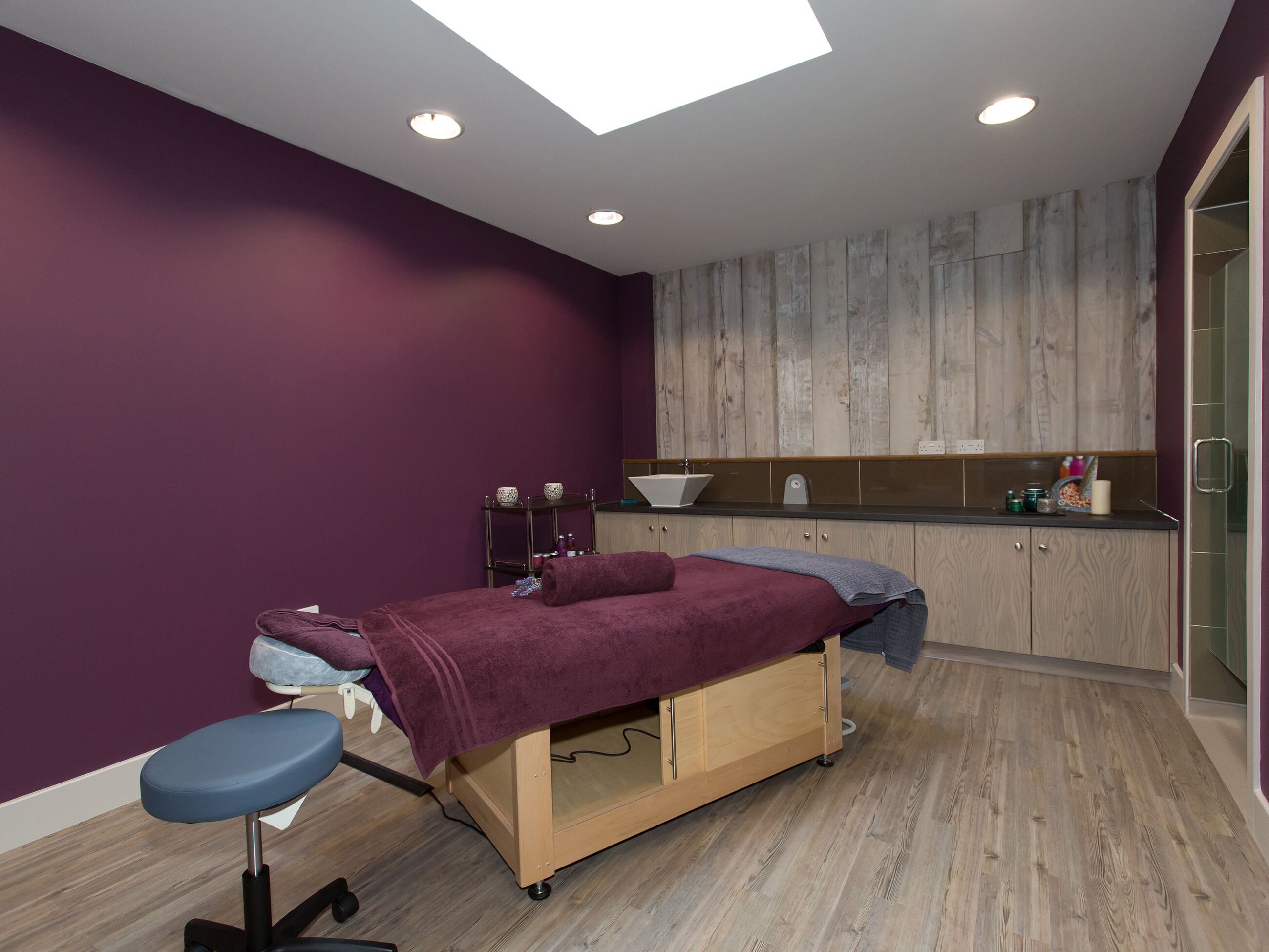 A room with a wooden floor, a bed with a purple blanket, a small table with a bowl, a mirror, and a wooden cabinet.