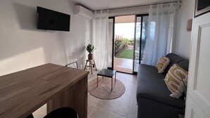 A modern living room with a wooden dining table, a sofa with cushions, a television mounted on the wall, and a sliding glass door leading to an outdoor area. - Apartment for 4 with sea view, pool, and pkg (Cannes)