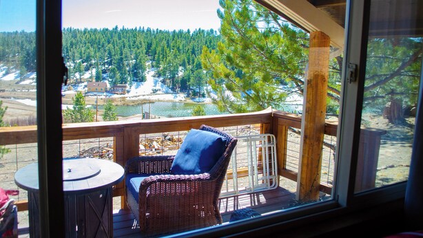Una terraza de madera con una silla de mimbre y una mesita, con vistas a un área boscosa con un lago y edificios a lo lejos.