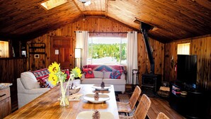 Interior acogedor de una cabaña con techo de madera, una mesa de comedor decorada con piñas, un sofá con almohadones coloridos y una chimenea.