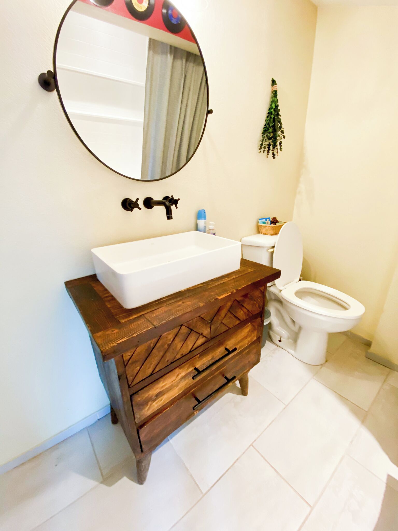 A bathroom with a white sink, a wooden vanity, a round mirror, a toilet, and a small plant on the wall.
