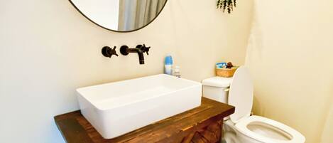A bathroom with a white sink, a wooden vanity, a round mirror, a toilet, and a small plant on the wall.