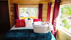 Habitación de cabaña de madera con cama, cortinas rojas y ventana con vistas a la vegetación.