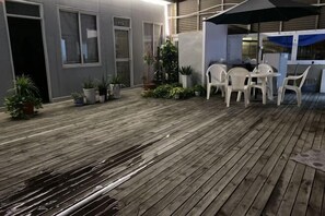 Zona de pati exterior amb paviment de fusta, cadires de plàstic blanc i plantes en testos.