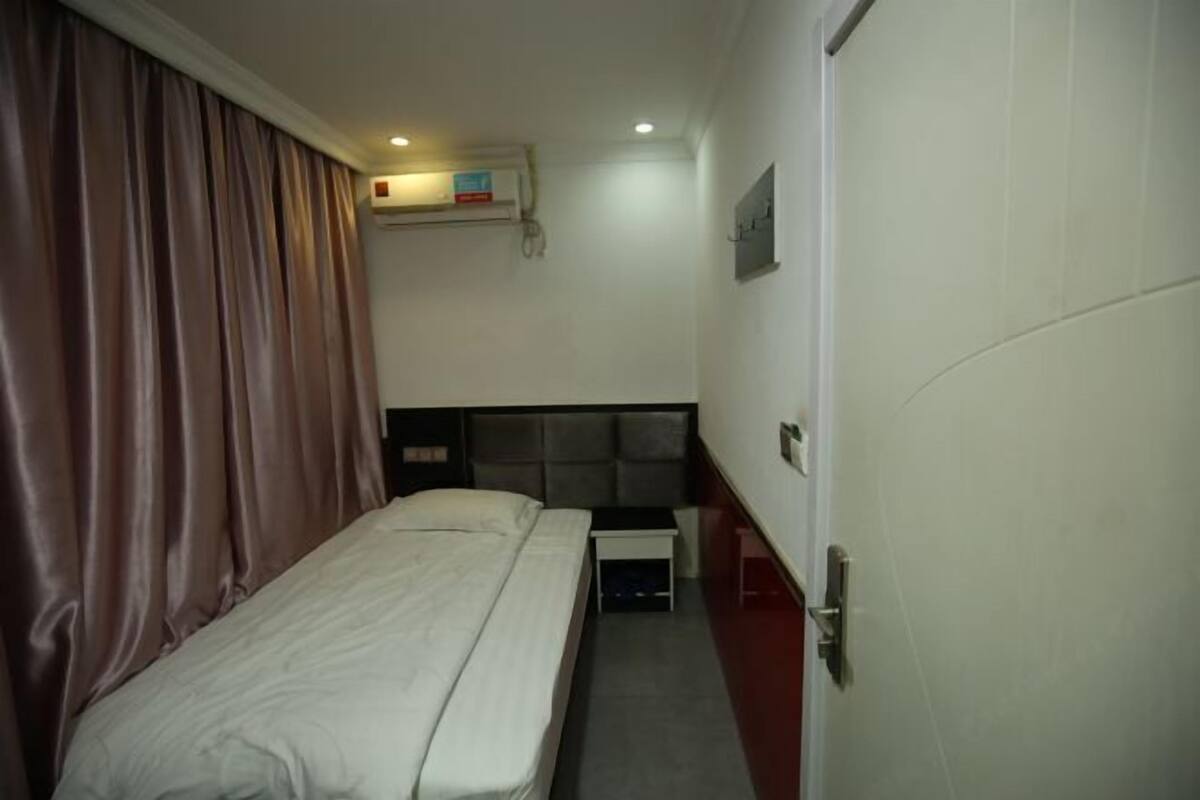 Habitación de hotel con cama, mesita de noche, un armario pequeño, aire acondicionado instalado en la pared y una puerta.