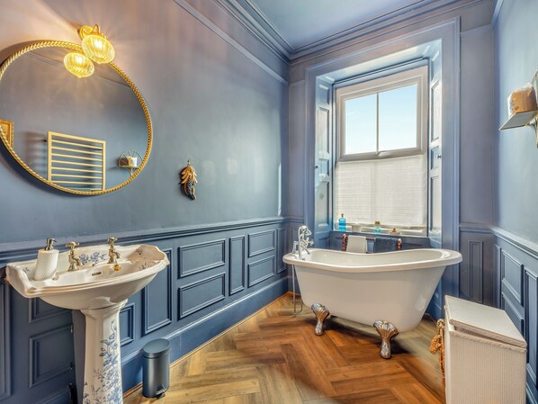 A bathroom with a freestanding bathtub, a pedestal sink, and a round mirror with a gold frame.