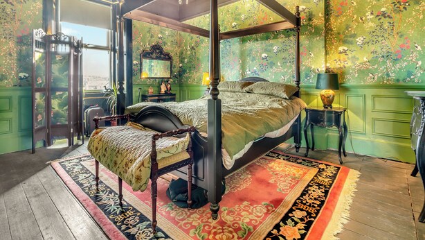 Een hemelbed met baldakijn, groene bloemenbehang en een tapijt met een rood-zwarte print.