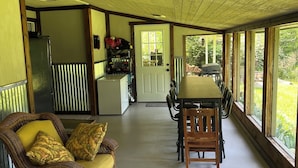Ein geräumiges Zimmer mit Esstisch und Stühlen, einem Korbsessel mit Kissen, einem Kühlschrank und Blick durch große Fenster.