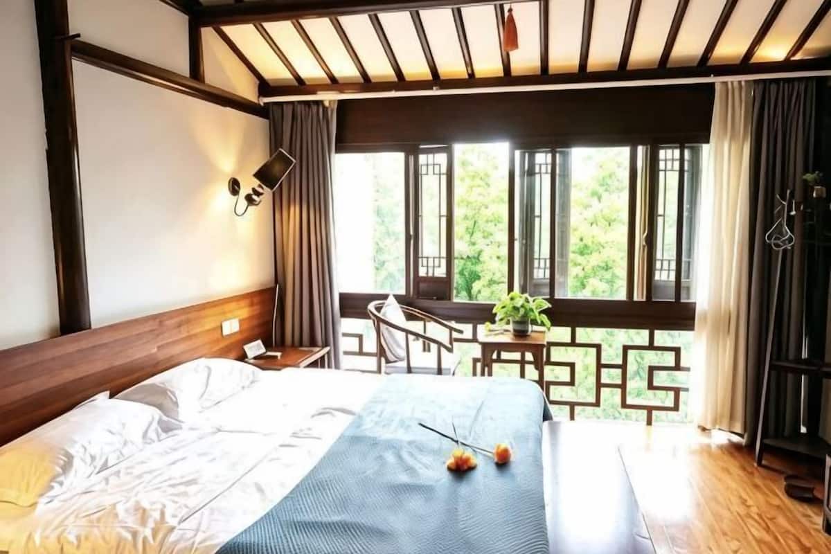 Un dormitorio con cama, escritorio, silla, ventana con cortinas y vista a la vegetación.