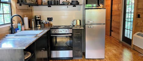 A compact kitchen with a stove, oven, microwave, and refrigerator. There is a sink with a faucet, and a window with blinds.