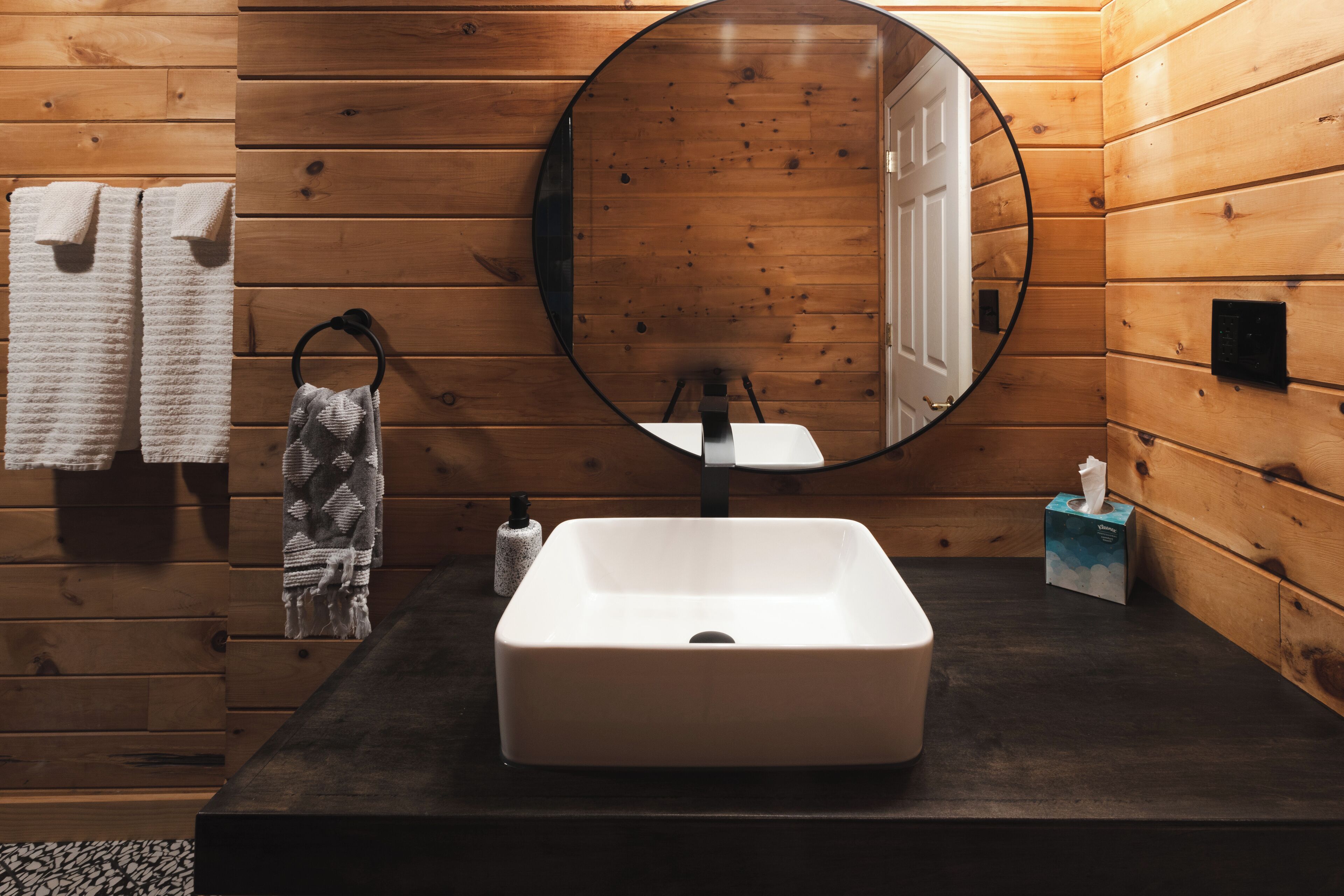 A bathroom with wooden walls, a round mirror, a white sink, and towels.
