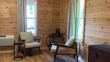 A cozy room with wooden walls, a ceiling fan, two windows with curtains, a coffee table, a chair, a sofa, a rug, and a dresser.