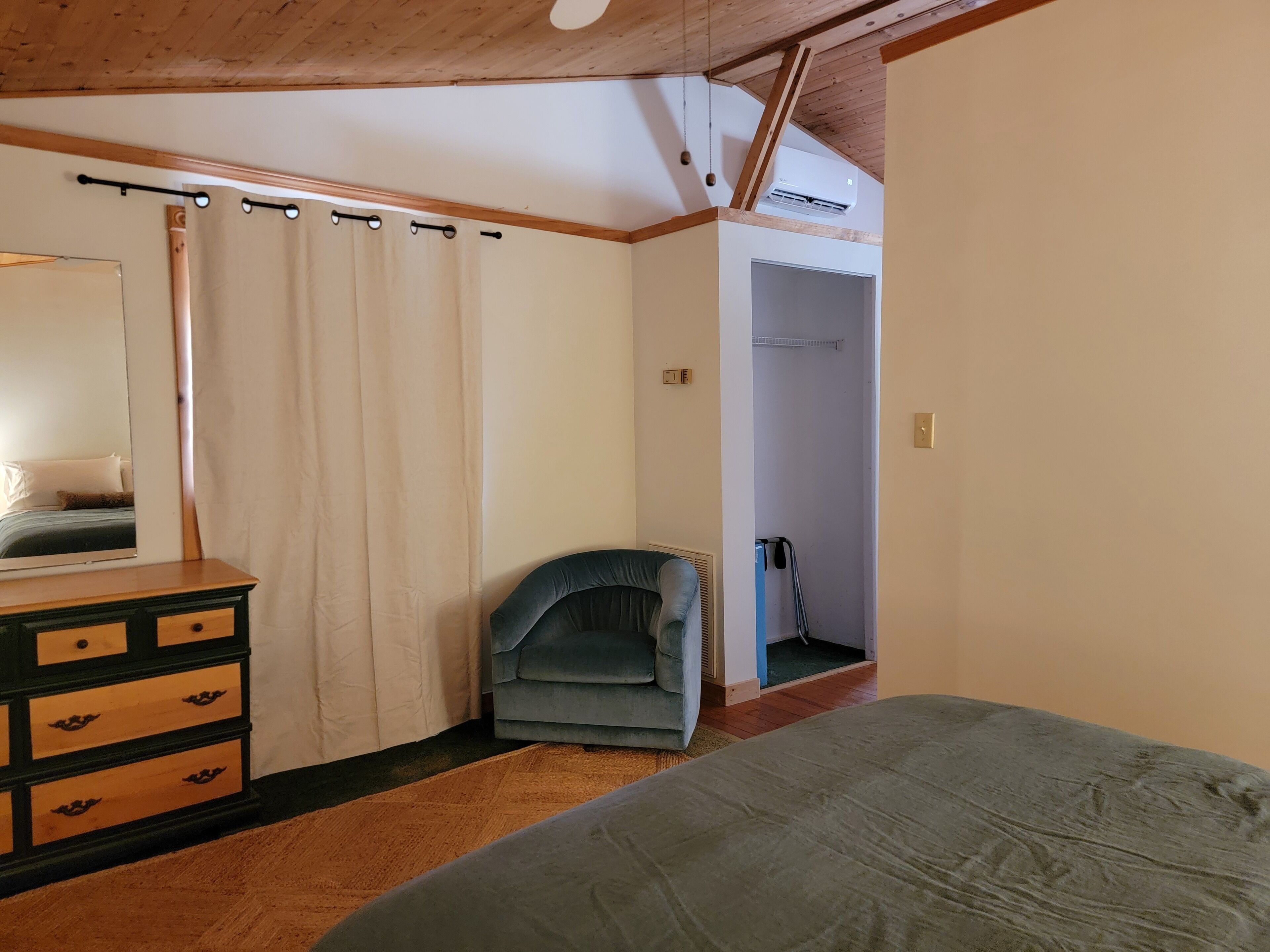 A bedroom with a bed, a dresser, a chair, a mirror, a closet, and an air conditioning unit.