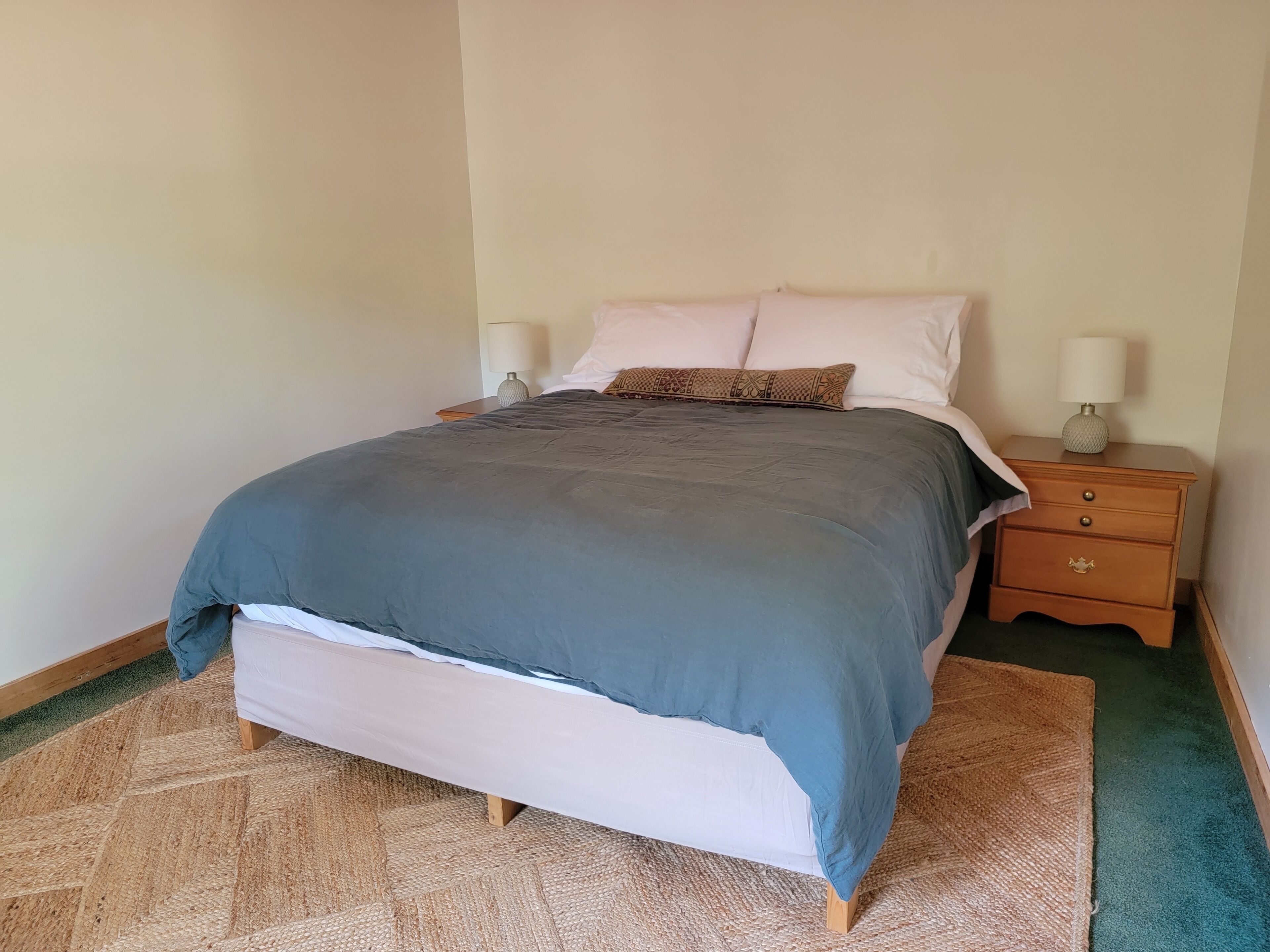 Una cama bien tendida con edredón gris y blanco, dos lámparas de noche y una mesita de noche de madera con cajones.