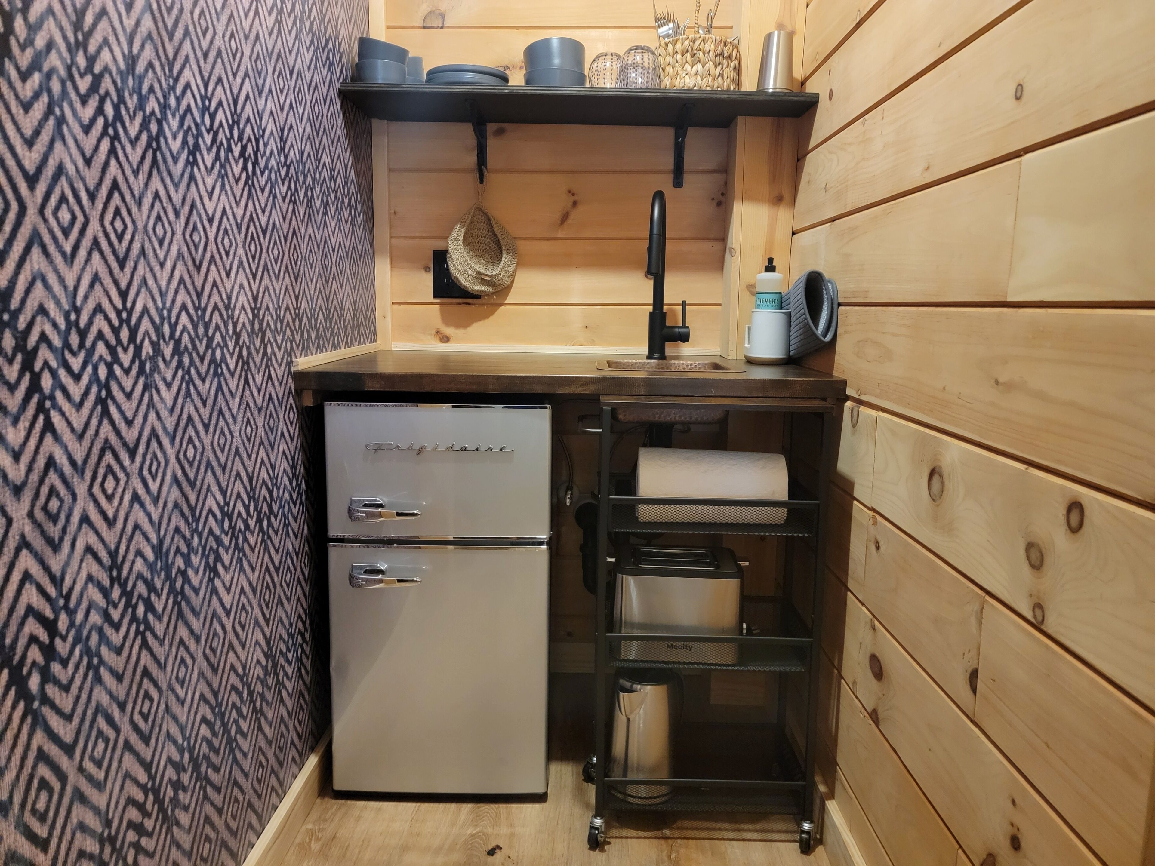 A compact kitchenette with a small refrigerator, a sink, and a metal rack for storage.