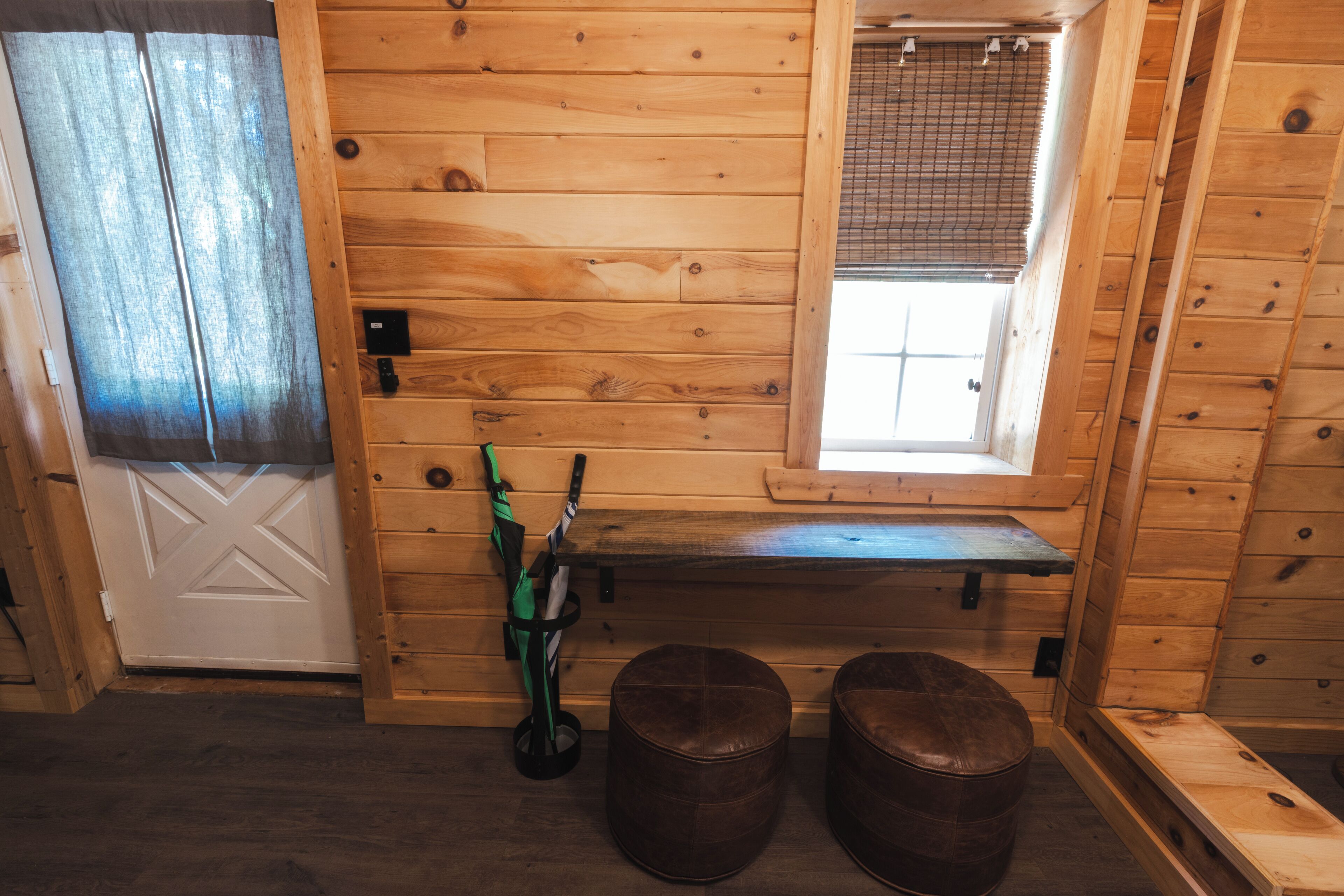 A wooden cabin interior with a bench, two ottomans, a door, and a window with a view.