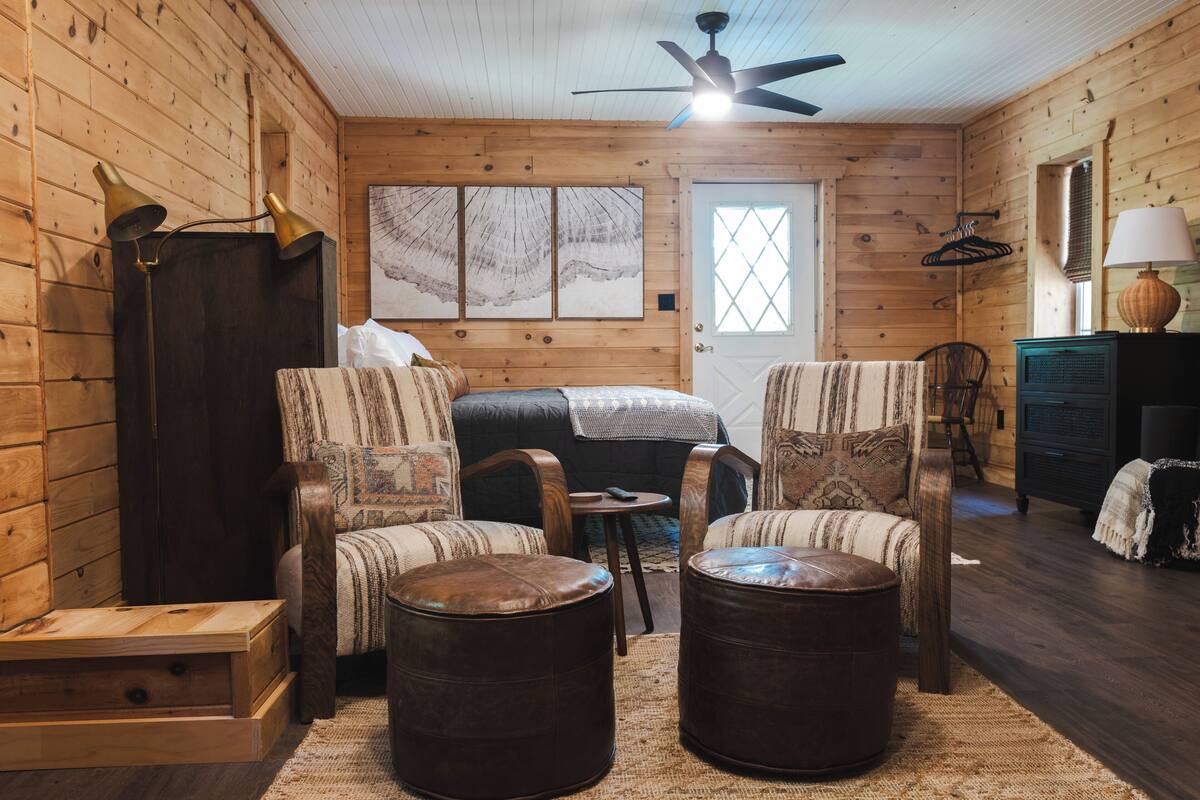 A cozy log cabin room with a bed, two armchairs, a small table, and a ceiling fan.