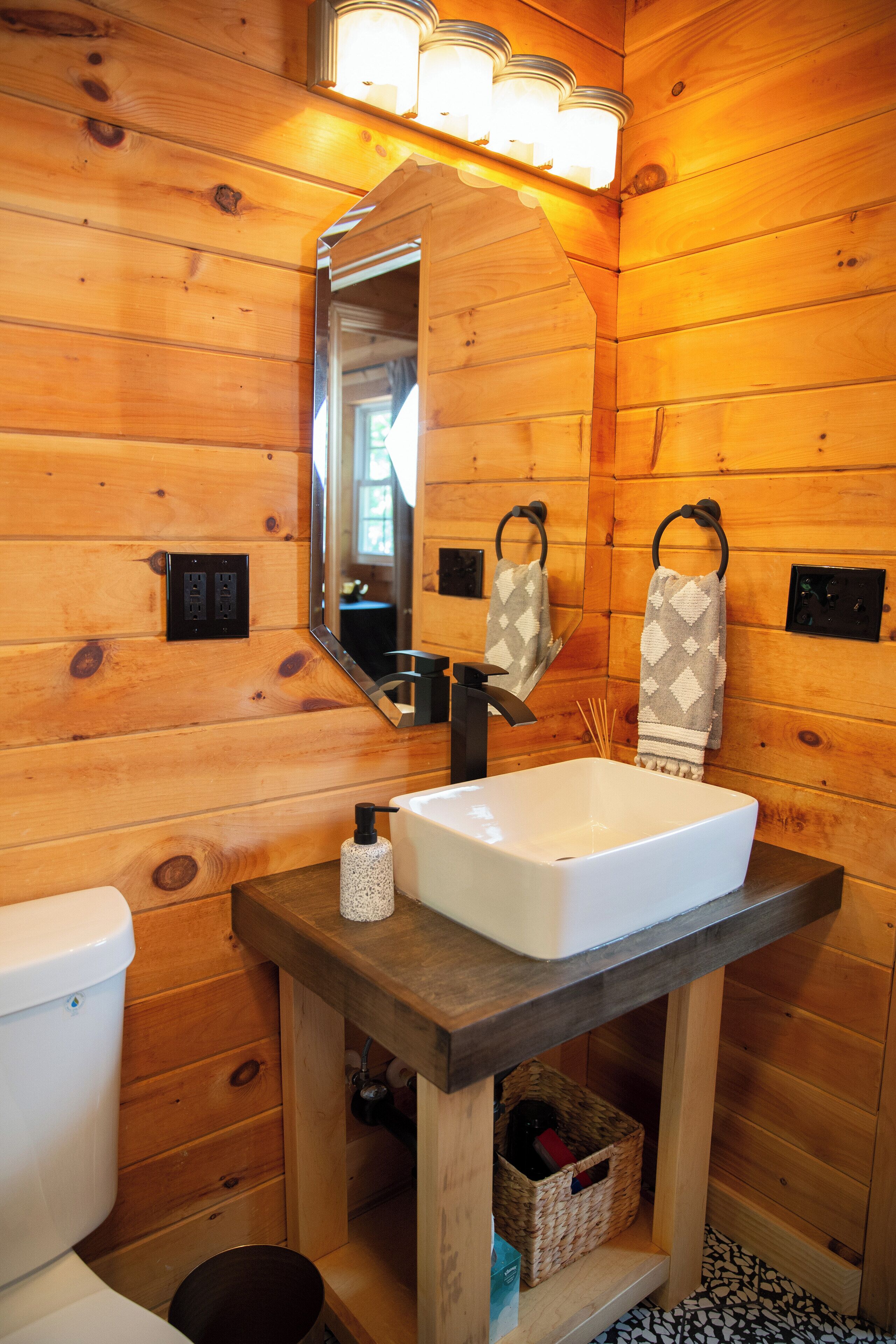 Une salle de bain rustique, avec des murs en bois, un lavabo blanc, un miroir et deux serviettes suspendues à des crochets.