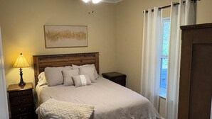 Ein Schlafzimmer mit einem Holz-Kopfteil, einem Bett mit vielen Kissen, einem Nachttisch mit Lampe und einem Fenster mit Vorhängen.