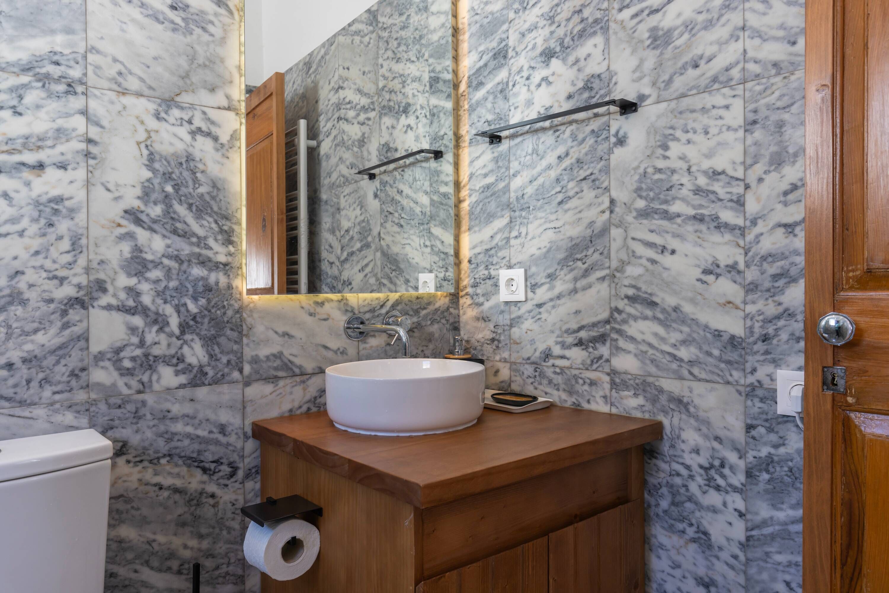 A bathroom with a marble backsplash, a wooden vanity, a white sink, and a towel rack.