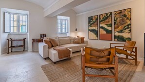 A spacious living room with a white sofa, a wooden coffee table, and a leather armchair. There are two windows with open shutters, a lamp, and three framed artworks on the wall.