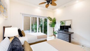 A modern bedroom with a bed, bedside table, lamp, TV, and a large window with a view of palm trees. - Tamarindo Beach Condo – 2BR Steps from Sea. (Tamarindo)