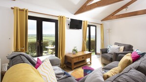 A living room with a sofa, a coffee table, and a TV. There are large windows with curtains, a wooden ceiling, and a view of the countryside. - *3 Bed Bungalow*Near Portreath Beach*Hot Tub*Free Parking & WiFi* (Redruth)
