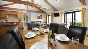 A dining area with a wooden table set for four, surrounded by black leather chairs. A kitchen with wooden cabinets and a sink is visible in the background. - *3 Bed Bungalow*Near Portreath Beach*Hot Tub*Free Parking & WiFi* (Redruth)