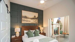 Une chambre à coucher bien aménagée, avec un grand lit, deux tables de chevet avec des lampes, un tableau au mur et une cheminée dans la pièce adjacente.