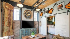 A rustic living room with exposed brick walls, a large flat-screen TV, a wooden coffee table, and a leather couch. - Industrial Design Apt | 10 Min to Downtown (Nashville)
