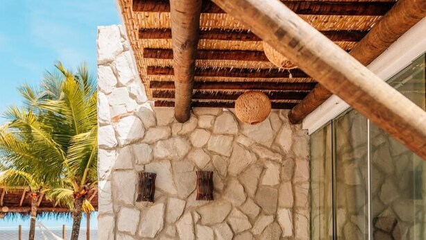 Une terrasse avec un mur en pierre, deux fauteuils en osier et une porte vitrée donnant sur la plage.