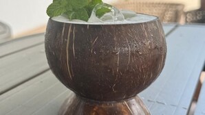 Une boisson à base de noix de coco sur une table en bois, avec un arrière-plan flou représentant un paysage extérieur.