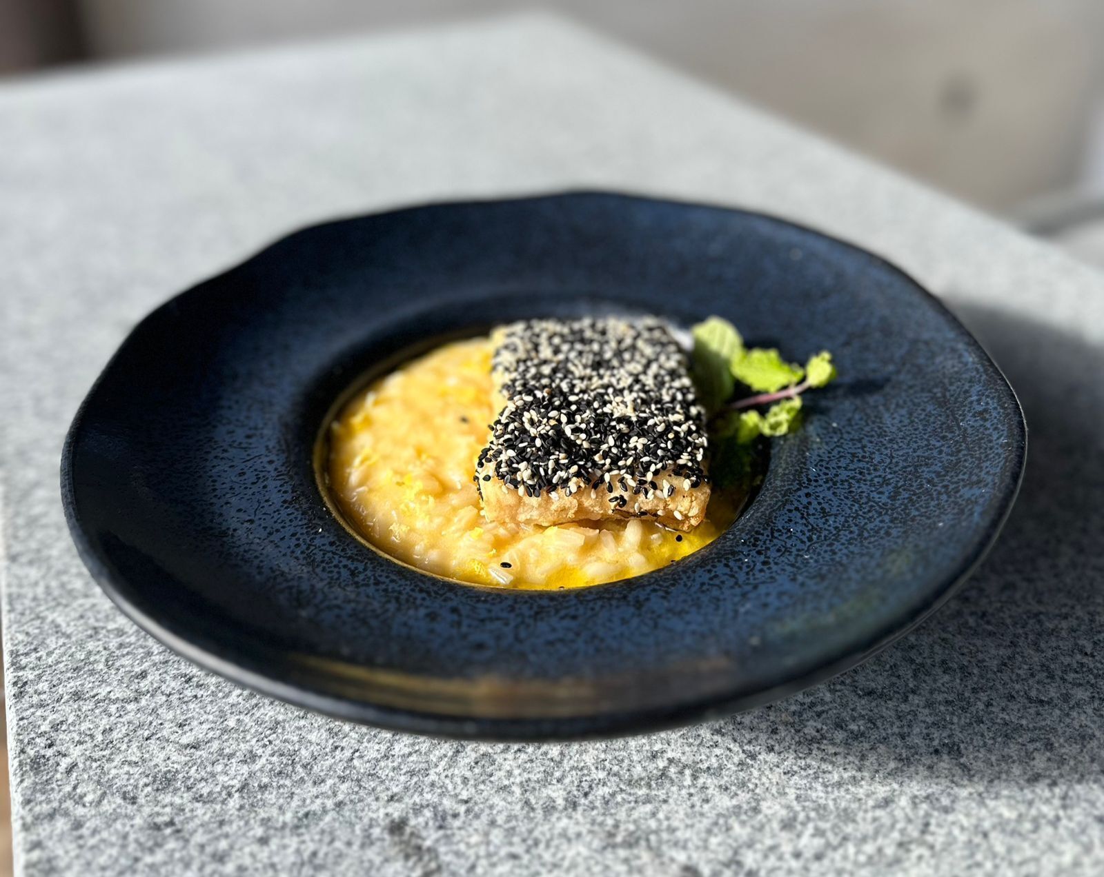 A dish with a fish fillet topped with sesame seeds, served on a dark plate.