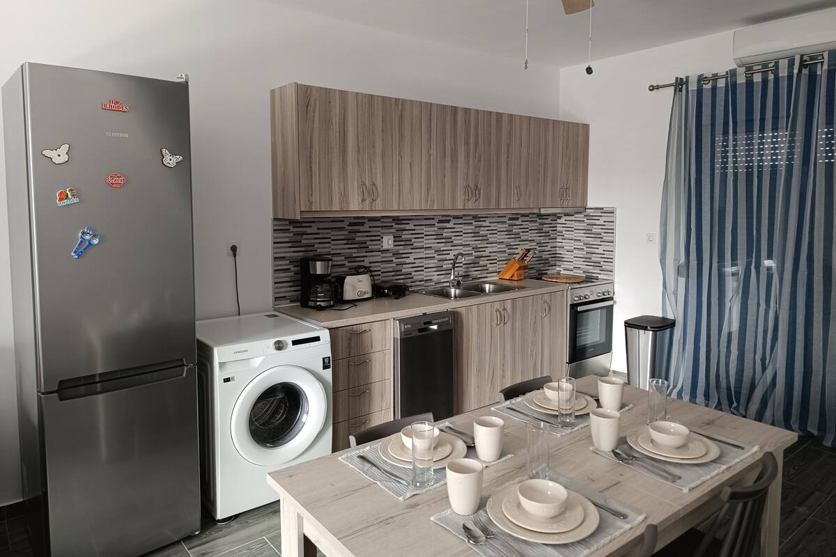 A modern kitchen with a stainless steel refrigerator, a washing machine, wooden cabinets, a coffee maker, and a dining table set for four.