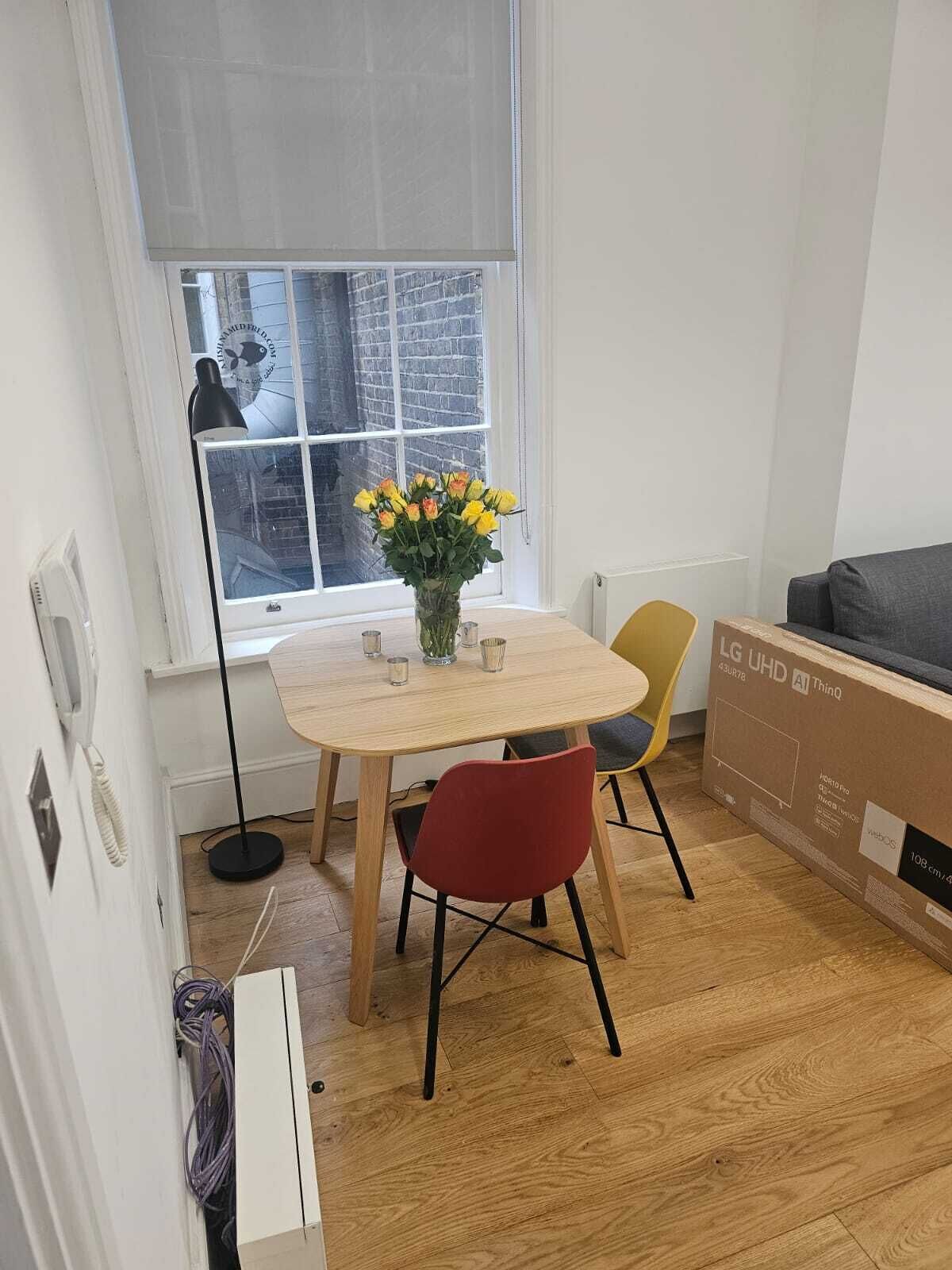 Une petite pièce moderne avec une table à manger en bois, deux chaises, un vase de fleurs, une fenêtre donnant sur un bâtiment en briques et un grand carton posé sur le sol.